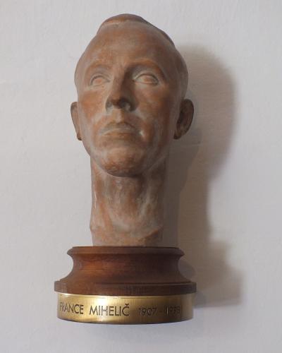 A terracotta bust of a man mounted on a wooden base with a brass plaque reading "FRANCE MIHELIČ 1907 - 1998" against a plain white wall.