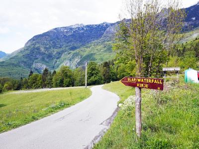 A wooden directional signpost on a grassy roadside verge pointing towards "SLAP/WATERFALL VIRJE" with a distance marker of 30, set against a backdrop of a winding paved road and green mountains.