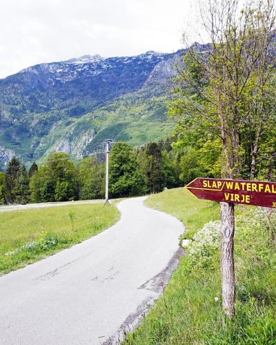 A wooden directional signpost on a grassy roadside verge pointing towards "SLAP/WATERFALL VIRJE" with a distance marker of 30, set against a backdrop of a winding paved road and green mountains.