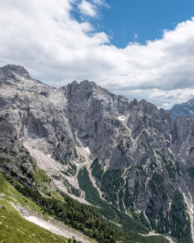 A high-angle landscape photograph showing a rugged, rocky mountain ridge with green alpine slopes in the foreground under a cloudy blue sky.