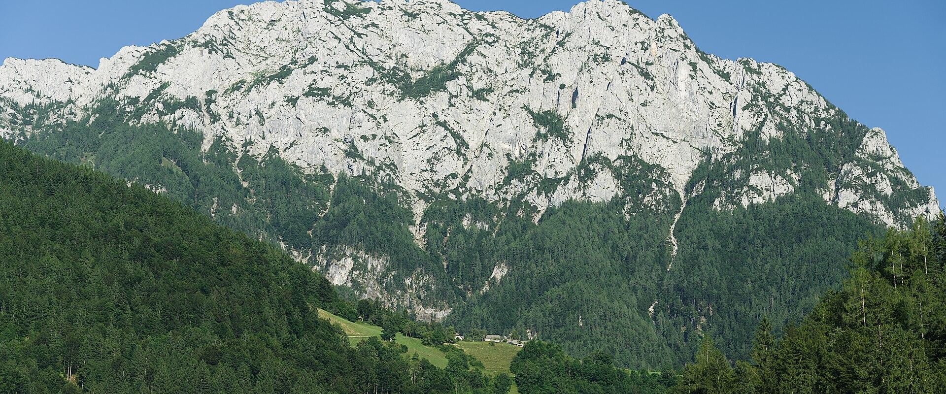 A large, rugged limestone mountain with a broad, rocky summit ridge rising above a dense green forest and a small settlement in the valley below.