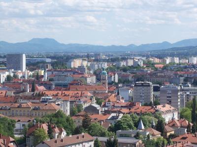 A panoramic view of a European city with red-tiled roofs, several church spires, and distant mountains under a cloudy sky.