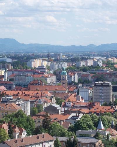 A panoramic view of a European city with red-tiled roofs, several church spires, and distant mountains under a cloudy sky.