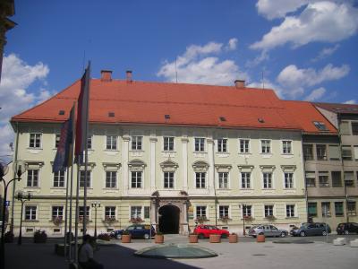 A large, pale yellow Baroque-style building with a red tiled roof and an arched gateway, situated in a paved public square with cars, flags, and a glass skylight structure in the foreground.