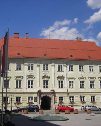 A large, pale yellow Baroque-style building with a red tiled roof and an arched gateway, situated in a paved public square with cars, flags, and a glass skylight structure in the foreground.