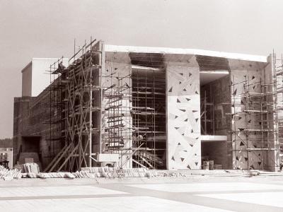 A black and white photograph showing a large modernist building under construction, covered in scaffolding, with distinctive geometric patterns on its facade and construction materials piled in the foreground.