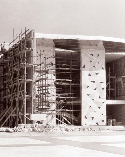 A black and white photograph showing a large modernist building under construction, covered in scaffolding, with distinctive geometric patterns on its facade and construction materials piled in the foreground.