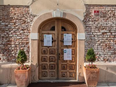 A close-up of an arched wooden double door set into a building facade with exposed brickwork on the sides and a beige stucco upper section, flanked by two potted plants and featuring posted notices on the doors and a red address plaque on the wall.