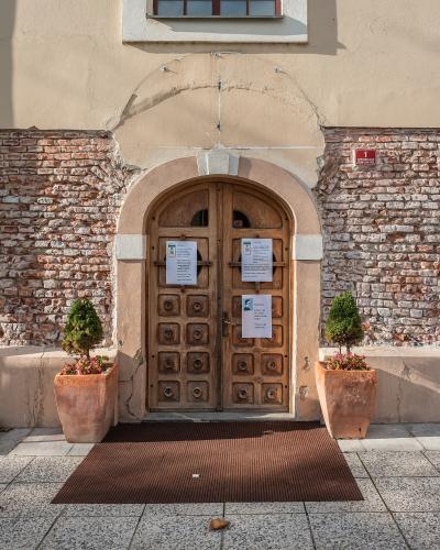 A close-up of an arched wooden double door set into a building facade with exposed brickwork on the sides and a beige stucco upper section, flanked by two potted plants and featuring posted notices on the doors and a red address plaque on the wall.
