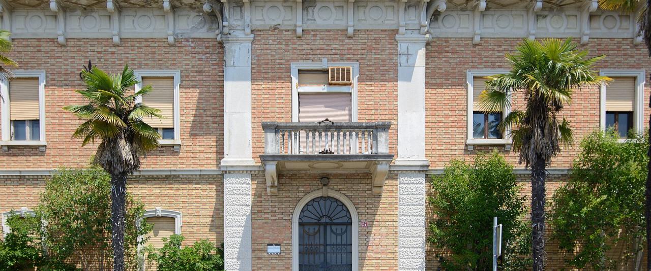 A two-story brick and stone building with a central balcony, arched doorway, palm trees, and decorative railings, identified by filename as "Piran office building Droga Portorož-4504".