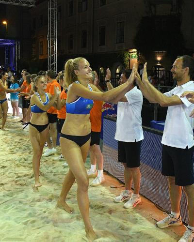 A group of female beach volleyball players in blue and black uniforms are high-fiving officials or coaches on a sand court at night, with spectators and event banners visible in the background.