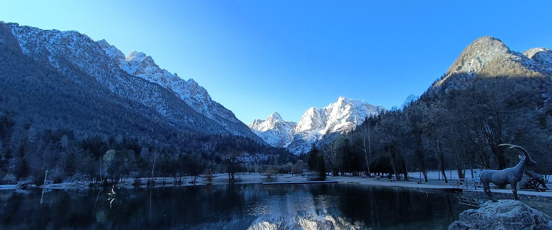 A frozen lake in a snowy alpine valley with a distinctive jagged mountain peak reflected in the open water in the foreground.