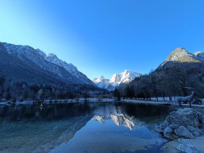 A frozen lake in a snowy alpine valley with a distinctive jagged mountain peak reflected in the open water in the foreground.