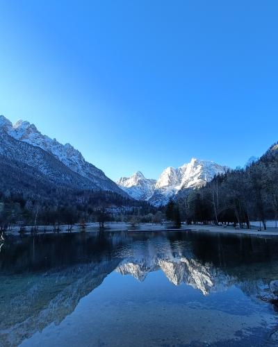 A frozen lake in a snowy alpine valley with a distinctive jagged mountain peak reflected in the open water in the foreground.