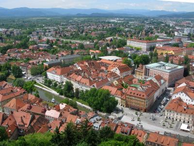Aerial view of Ljubljana’s city center, showing the Ljubljanica River, red-tiled roofs, and surrounding urban landscape with mountains in the background; no prominent signage or specific building labeled.