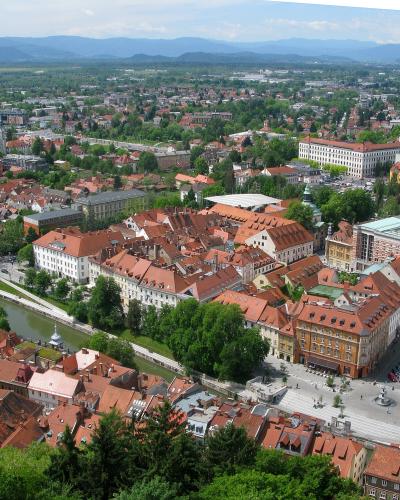 Aerial view of Ljubljana’s city center, showing the Ljubljanica River, red-tiled roofs, and surrounding urban landscape with mountains in the background; no prominent signage or specific building labeled.
